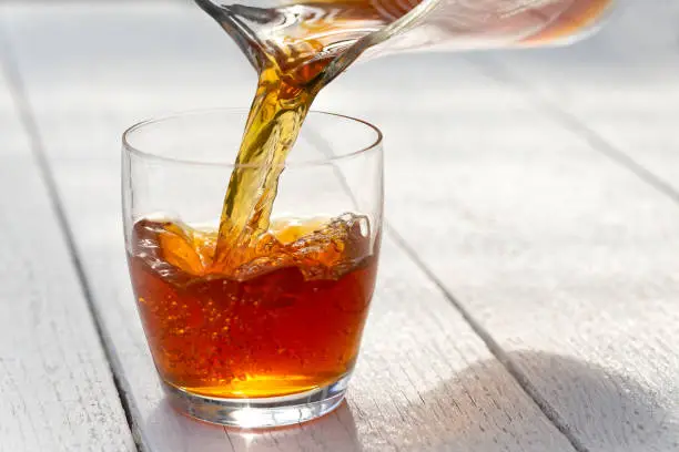 Pouring ice tea from a glass jug into a glass. White wood background.