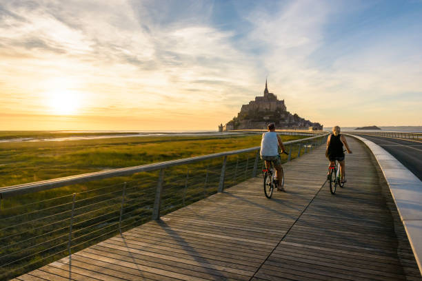 View of the Mont Saint-Michel tidal island in Normandy, France, at sunset with a couple biking to the town on the wooden jetty. Le Mont-Saint-Michel, France - June 24, 2018: A couple of tourists biking at sunset on the new jetty leading to the tidal island since 2014 as part of the dredging program of the bay. mont saint michel photos stock pictures, royalty-free photos & images