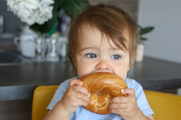 Portrait of adorable funny baby boy holding and biting big bagel sitting on yellow chair at home kitchen. Close-up. stock photo