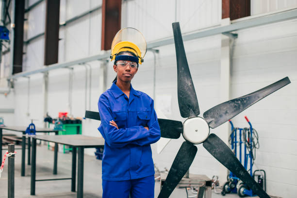 Portrait of a Young Woman in an Engineering Workshop A young woman stands and looks at the camera in an engineering workshop. She is wearing a protective mask and eyewear as well as blue coveralls. northeastern england photos stock pictures, royalty-free photos & images