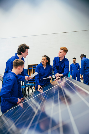 A group of engineering students stand next to some solar panels and work on a group project together. They are all wearing blue coveralls.