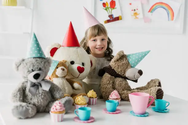 Photo of smiling birthday kid with teddy bears in cones having tea party with cupcakes at table