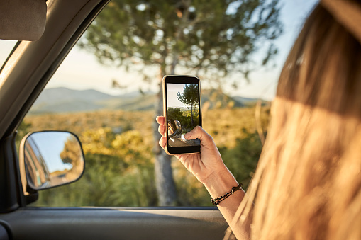 Cropped image of woman photographing nature from car window. mid adult female is capturing a perfect road trip moment. Focus is on her hand holding smart phone.