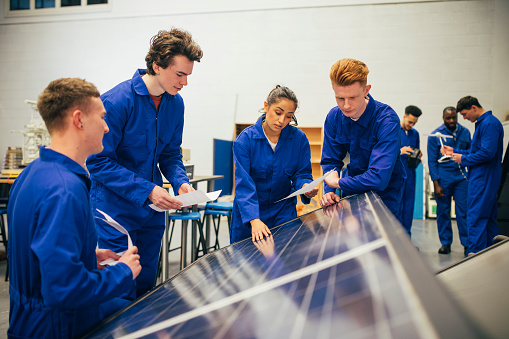 A group of engineering students stand over some solar panels and work on a group project together. They are all wearing blue coveralls.