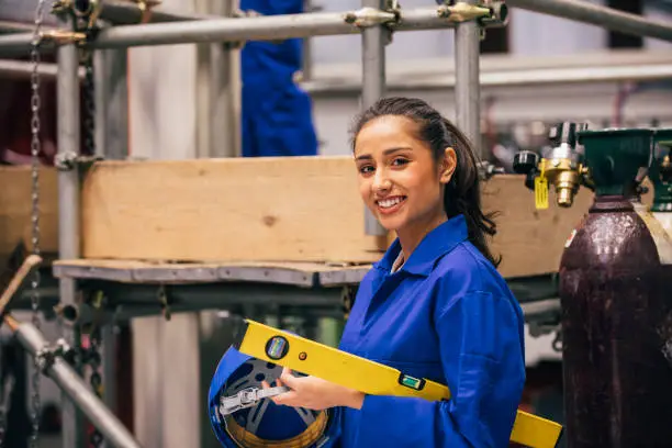 A young Indian woman looks at the camera as she stands in an engineering workshop and carries a spirit level and work helmet - she is also wearing blue coveralls.