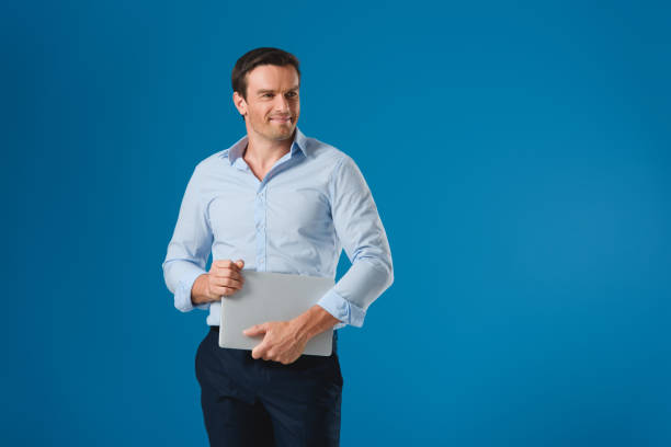 istock handsome smiling man holding laptop and looking away isolated on blue 999101072