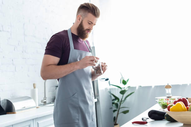 istock handsome bearded man in apron holding container with cinnamon sticks while cooking in kitchen 999101206