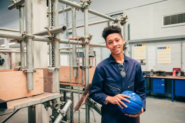 A mixed race young man looks at the camera as he stands in an engineering workshop and carries a work helmet - he is also wearing blue coveralls.