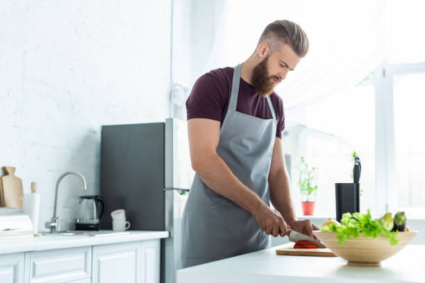 istock handsome bearded young man in apron cutting vegetables while cooking in kitchen 999104718