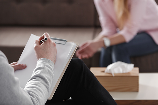 Primer plano de la mano del terapeuta escribir notas durante una sesión de consejería con una mujer sentada en un sofá en el fondo borroso. photo