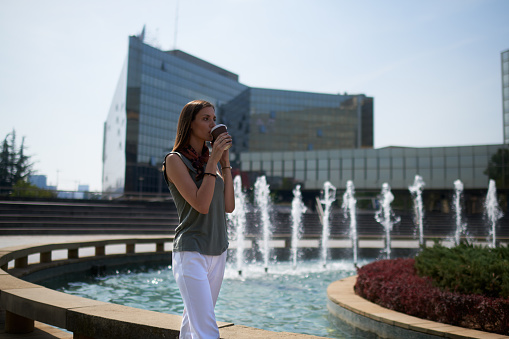 Young fashionable female in the city having coffee drink on the move. There is a fountain in the background.