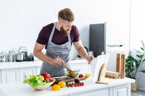 istock handsome bearded man in apron cooking vegetable salad and reading cookbook 999111674