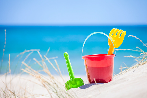 Horizontal shot of a sand bucket, pail and shovel taken on white sand dune against turquoise colored beach background. The objects are grouped at the left of an horizontal frame leaving useful copy space for text and/or logo at the center-right. Predominant colors are blue and red. DSRL outdoors photo taken with Canon EOS 5D Mk II and Canon EF 70-200mm f/2.8L IS II USM Telephoto Zoom Lens