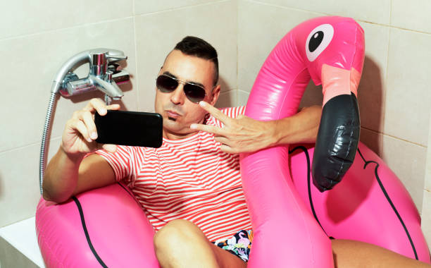 man with a swim ring taking a selfie indoors a young caucasian man wearing sunglasses, a swim suit and a swim ring in the shape of a pink flamingo in the bathtub of his bathroom, taking a self-portrait with his smartphone staycation stock pictures, royalty-free photos & images