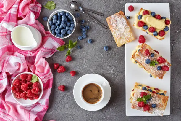 french dessert millefeuille of puff pastry and custard cream, raspberries, blueberries on a plate with cup of coffee, fresh cream and berries on a table, view from above, flat lay