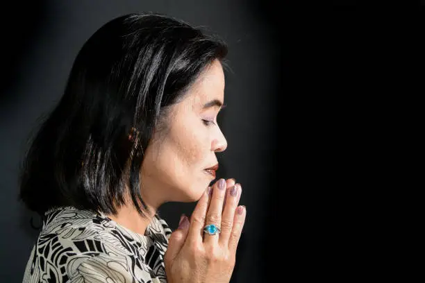 This horizontal image shows a profile of a mature adult Asian Filipino woman with hands clasped and praying. She is wearing a patterned brown blouse, has black hair and the image was shot in the studio against a black background.