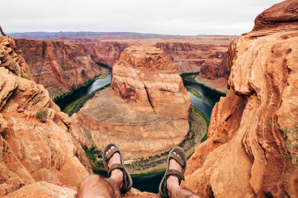 Personal perspective view of mature man sitting with feet hanging over edge of cliff at Horseshoe Bend canyon