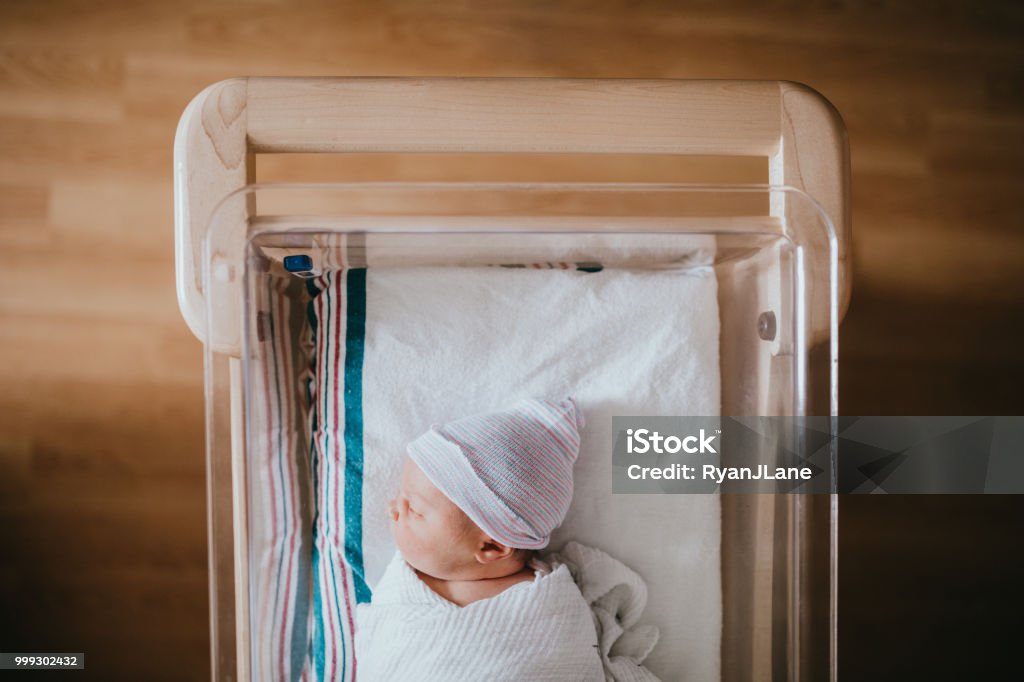 Newborn Baby Sleeping In Hospital Bassinet A baby just born at the hospital rests in a hospital bassinet crib, wrapped in a swaddle and wearing a beanie hat. Newborn Stock Photo
