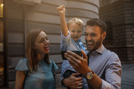 Family with one child outdoors on city street at night. They watching sport game on smart phone and walking in city centre