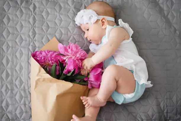 Happy newborn baby lying on the bed and playing with ping bouquet of pion flowers