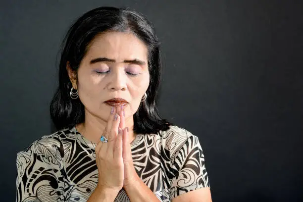 This horizontal image shows a mature adult Asian Filipino woman praying with her head bowed and eyes closed and hands together. She is wearing a brown patterned blouse, has black hair and the image was shot in the studio against a black background.