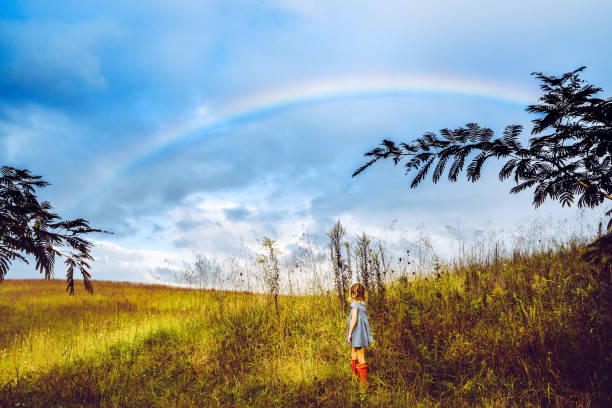Somwhere Over the rainbow... Adorable little girl in a blue dress and red boots in a vast nature field meadow area with a stunning rainbow in the sky. She is headed for adventure or maybe to find home? homeward stock pictures, royalty-free photos & images