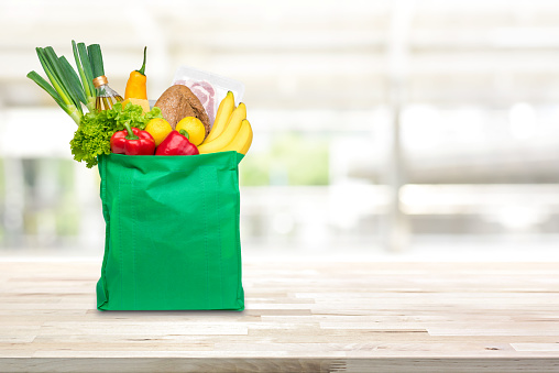 Food and groceries in green eco-friendly reusable shopping bag on wood table with blurred kitchen background
