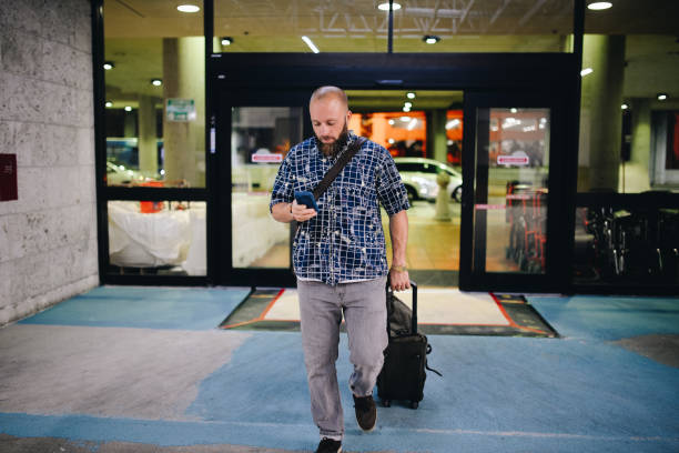 Airport hustle Man walks through the sliding glass doors of an airport, he pulls a suitcase and wears a carry on strap. He is dressed casually and has a beard, is looking at his phone, perhaps working from his phone. walking into glass door stock pictures, royalty-free photos & images