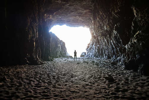 Woman silhouette standing by lighted exit in cave, daytime. Atmospheric snapshot in natural rock formations with sandy floors, tourist attraction