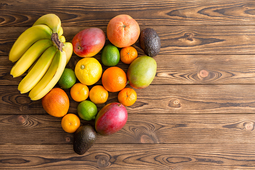 Heart-shaped still life of mixed healthy fresh tropical fruit with bananas, mango, orange, lemon, lime, avocado pear, clementine, mandarin on a wooden background with copy space