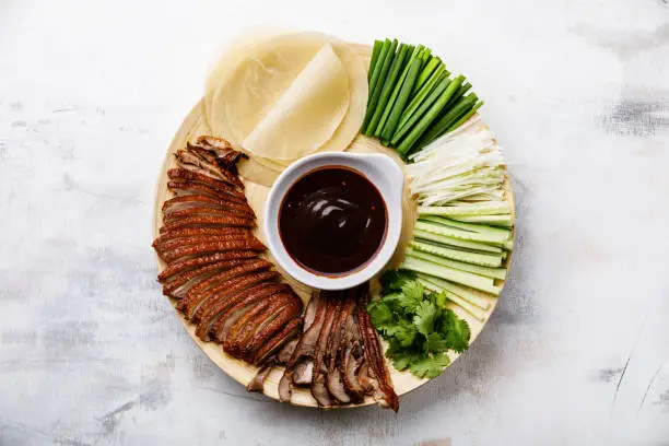 Sliced Peking Duck served with Hoysin sauce, fresh cucumber, onions, cilantro and roasted wheaten pancakes on white wooden background