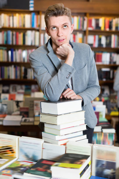 istock Pensive young man posing on stack of books in bookstore 999845710