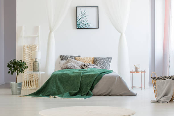 istock Green blanket thrown on double bed with many pillows and grey sheets standing in white hotel room interior with simple poster on the wall, fresh plant standing on the floor and glass lamp on bedside table 999868326