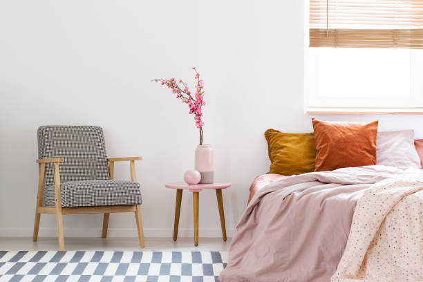 istock Flowers on table between patterned armchair and pink bed in bedroom interior with window. Real photo 999868778