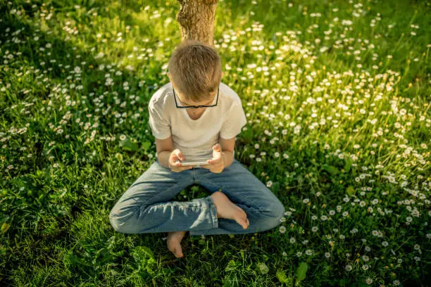 A child with smart phone sitting on the grass. Relax and texting with friends sitting outdoor barefoot