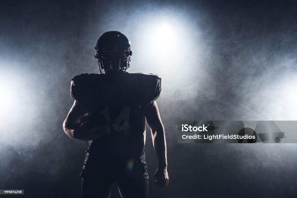 dark silhouette of equipped american football player with ball against white smoke American Football - Sport Stock Photo