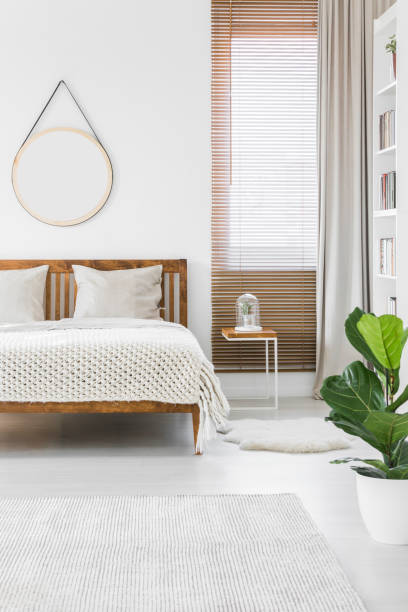 istock Knit blanket placed on wooden double bed in bright bedroom interior with empty round poster on the wall, window with blinds and green plant 999876556