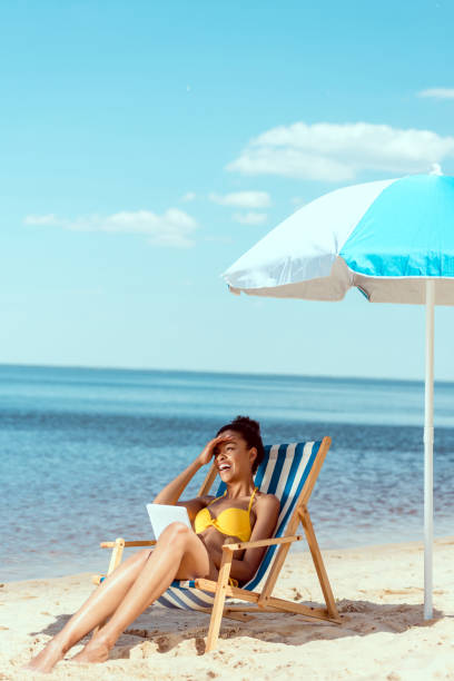 istock laughing african american woman relaxing on deck chair and using digital tablet under beach umbrella in front of sea 999876908
