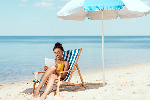 african american woman relaxing on deck chair and using digital tablet under beach umbrella in front of sea