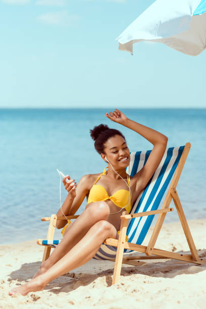 happy african american woman laying on deck chair and listening music in earphones with smartphone under beach umbrella in front of sea happy african american woman laying on deck chair and listening music in earphones with smartphone under beach umbrella in front of sea black women in bathing suits stock pictures, royalty-free photos & images