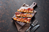 Fried bacon on wooden cutting board with fork and knife. Top view, isolated on black background.