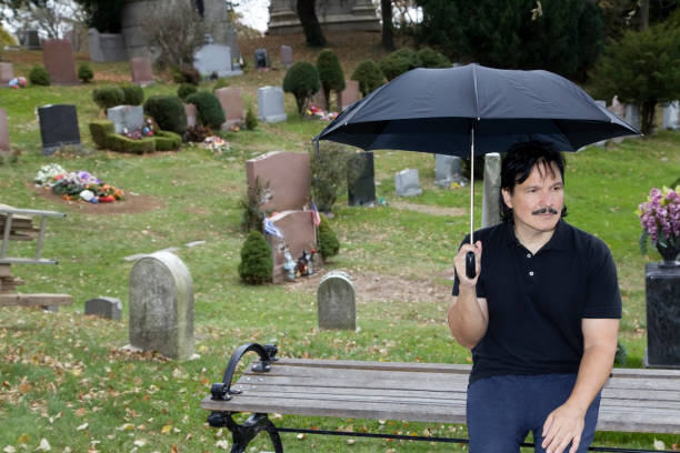 istock Latino man sits with umbrella on bench in cemetery 999922252