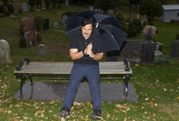 istock Man holding umbrella while seated in a cemetery 999922266