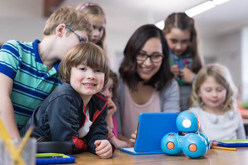 An ethnic female teacher is surrounded by her 1st grade students as she shows them how to program a robot using a digital tablet. One of the boys is smiling at the camera.