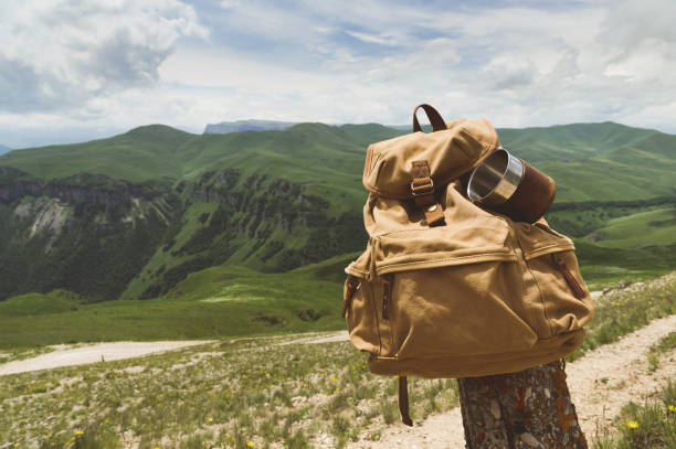 istock Hipster yellow vintage backpack with a mug fixed on it with a mug close-up front view. Traveler's travel bag in the background of a mountain landscape 999958286
