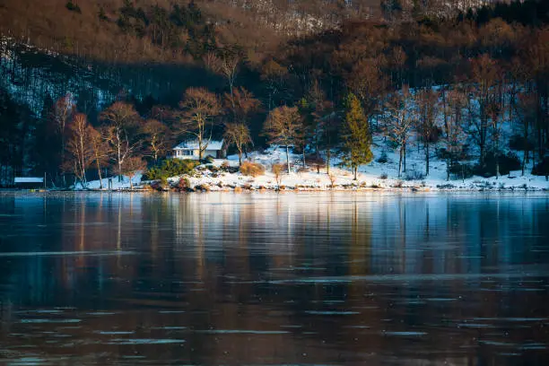 Evening view over the frozen Obersee of lake Rursee with a house by the shore in winter in the Eifel, Germany.