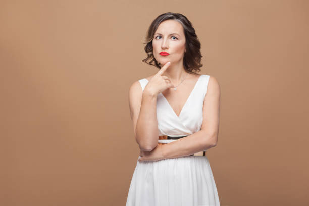 Beautiful adult woman thinking and puzzled Beautiful adult woman thinking and puzzled. Emotional expressing woman in white dress, red lips and dark curly hairstyle. Studio shot, indoor, isolated on beige or light brown background one mature woman only stock pictures, royalty-free photos & images