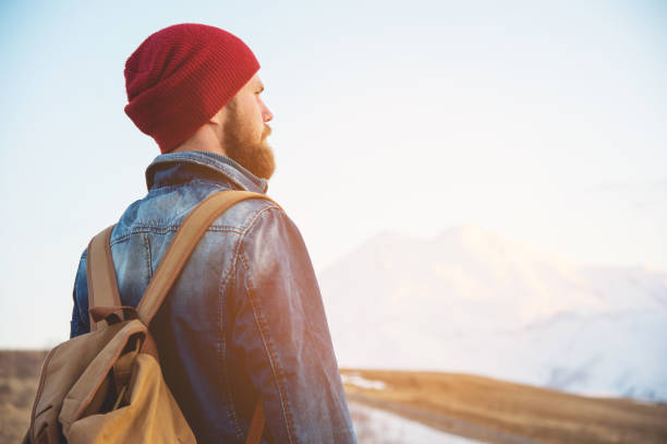 istock Bearded tourist hipster man in a hat with a backpack stand back on a roadside bump and watching the sunset against the background of a snow capped mountain 999963660