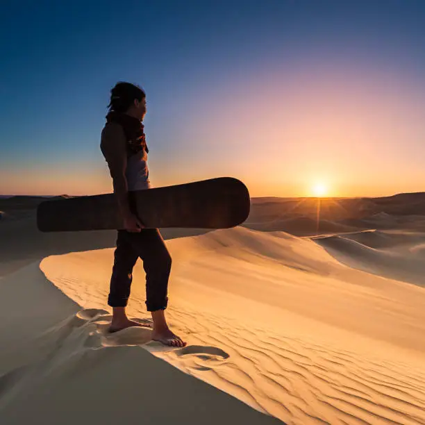 Young female sandboarding in The Sahara Desert during the sunset. She is standing on a sand dune, holding sandboard and watching the sunset over desert. The Sahara Desert is the world's largest hot desert with the biggest sand dunes.