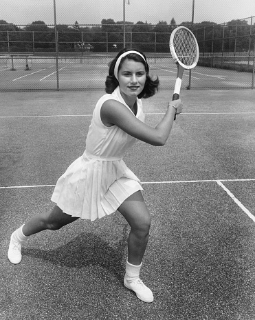 Fashionable woman in white clothing with tennis racket posing at tennis net on court. Sports Fashion